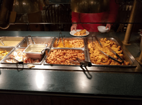 A buffet with various dishes, including shrimp and fried items, with a person serving food on a plate.