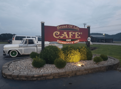 Sign for "Bread of Life Café" with established date 1988, surrounded by shrubs and a vintage truck parked nearby.
