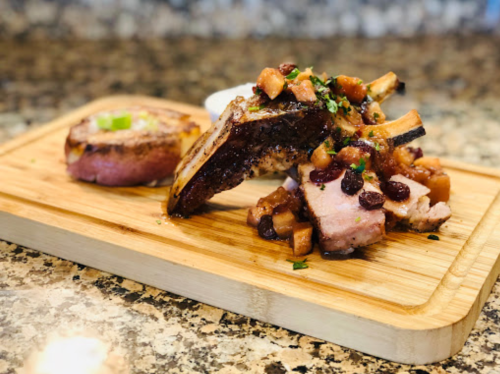 A wooden platter with grilled meat, apple chutney, and a side of sauce, set on a granite countertop.