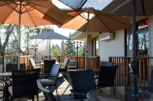 Outdoor patio with tables and chairs, shaded by large umbrellas, surrounded by trees and a building in the background.