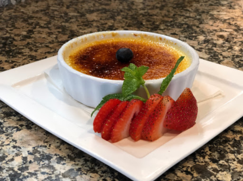 A creamy crème brûlée topped with a blueberry, served with sliced strawberries and mint on a white plate.