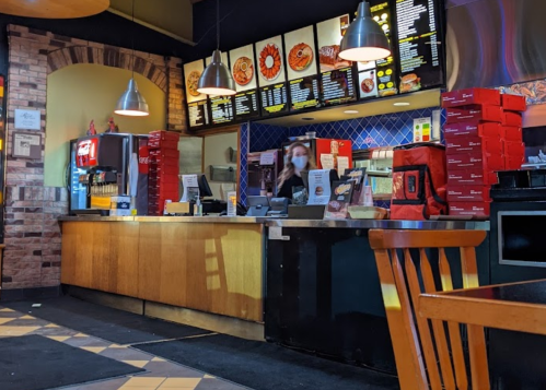 A restaurant counter with a menu board, a masked employee, and stacked pizza boxes in a cozy dining area.
