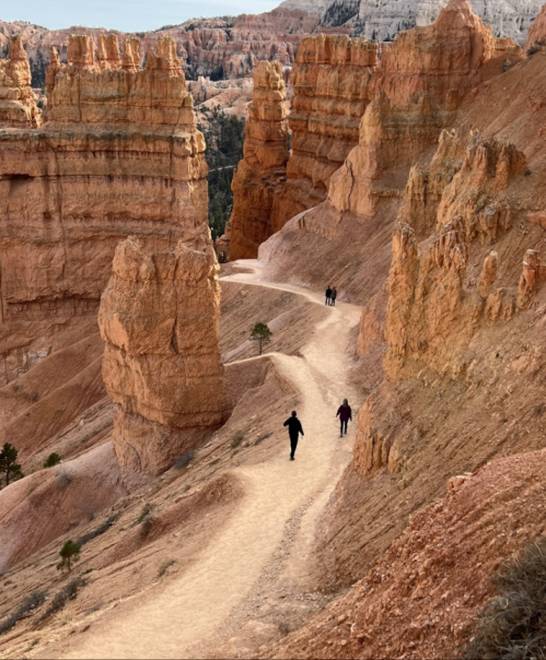 A winding path through orange rock formations in a canyon, with two hikers in the distance.