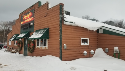 A log cabin-style restaurant with a snow-covered roof and festive decorations, surrounded by winter scenery.