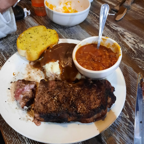 A plate with a steak, mashed potatoes with gravy, baked beans, and a slice of garlic bread.