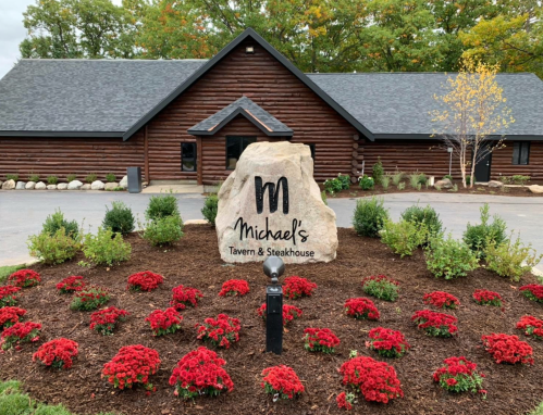 Sign for Michael's Tavern & Steakhouse in front of a log cabin building, surrounded by red flowers and greenery.