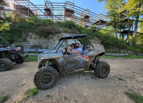 Two people sit in a muddy ATV parked on a dirt path, with a wooden structure and trees in the background.