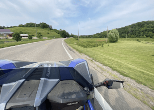 A view from a vehicle on a winding road, surrounded by green fields and hills under a blue sky.