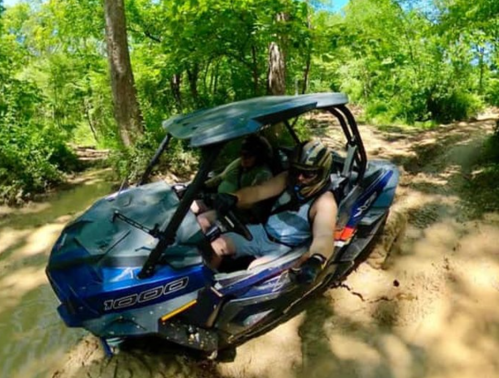 Two people riding in a blue off-road vehicle on a muddy trail surrounded by green trees.