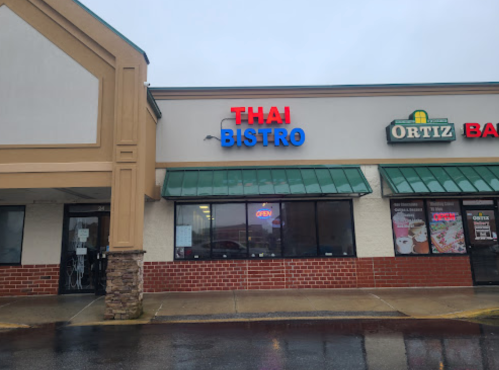 Exterior of a Thai Bistro restaurant with a bright "OPEN" sign, located in a shopping plaza on a rainy day.