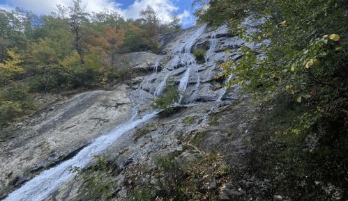 A cascading waterfall flows down a rocky cliff, surrounded by autumn foliage and a blue sky.