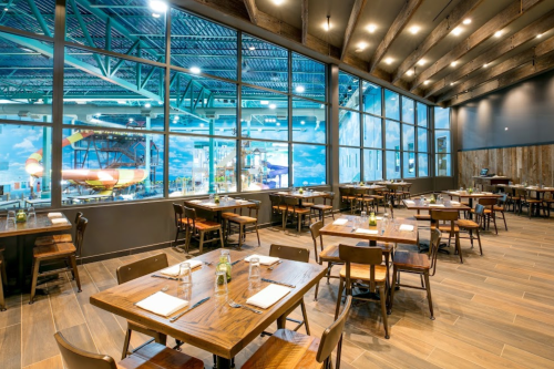 Modern restaurant interior with wooden tables, chairs, and large windows showcasing an industrial view outside.