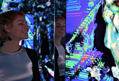 A smiling person stands beside a mirrored wall displaying vibrant, abstract digital art.