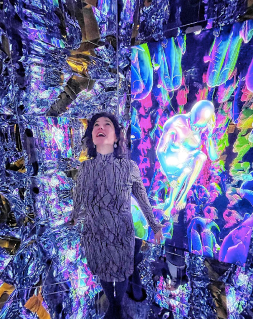 A woman stands in a mirrored room filled with colorful, abstract lights and a glowing, reflective figure.