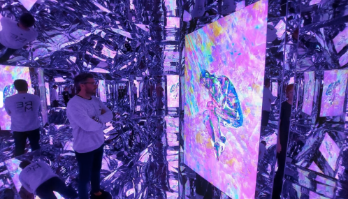 A person stands in a mirrored room, gazing at a colorful abstract artwork surrounded by reflective surfaces.