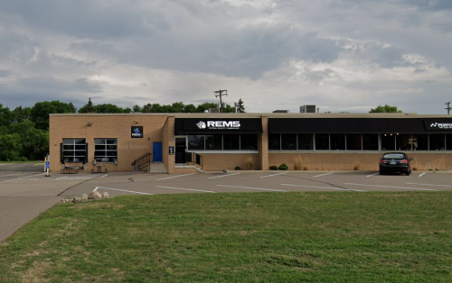 A brick building with large windows and a sign reading "REMS," surrounded by a grassy area and cloudy sky.