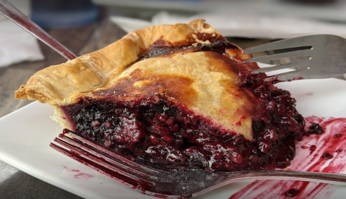 A slice of berry pie on a plate, with two forks partially digging into the filling, showcasing its rich, dark fruit interior.