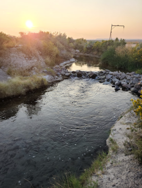 A serene river flows through a grassy landscape at sunset, with gentle ripples and a warm glow in the sky.