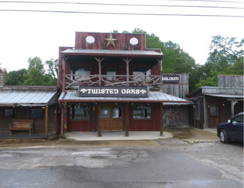 A rustic wooden building with a sign reading "Twisted Oaks" and a saloon, surrounded by greenery.