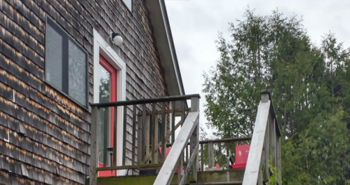 A wooden building with a red door and stairs leading up, surrounded by trees under a cloudy sky.