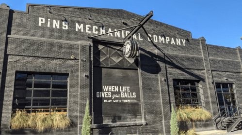Exterior of Pins Mechanical Company, featuring a black brick facade and a playful sign that reads, "When life gives you balls, play with them."