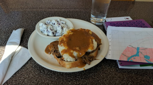 A plate with pulled meat on bread, mashed potatoes topped with gravy, a side of coleslaw, and a glass of water.