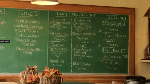 Chalkboard menu displaying daily specials, sides, salads, soups, and picnic basket options in a cozy restaurant setting.