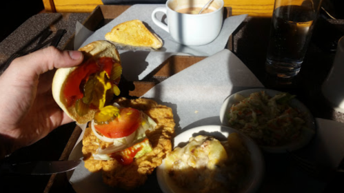 A hand holds a sandwich with toppings, alongside a cup of coffee, toast, and a bowl of coleslaw on a table.