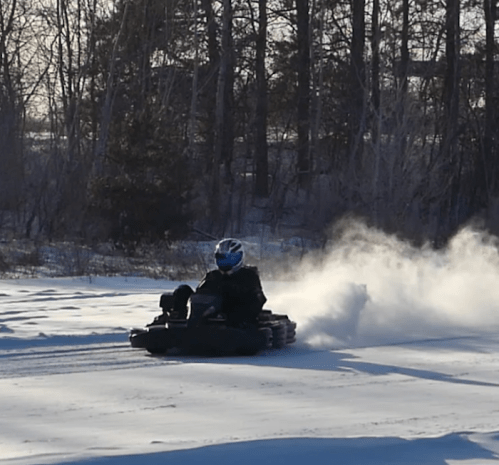 A person in a blue helmet drives a go-kart on snowy terrain, leaving a trail of snow and smoke behind.