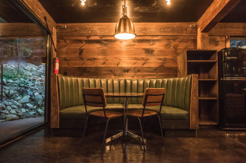 Cozy wooden interior with a green booth, a round table, and two chairs, illuminated by a pendant light.