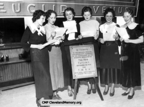 Six women in vintage clothing sing together in front of a sign honoring the New Taylor, September 1944.