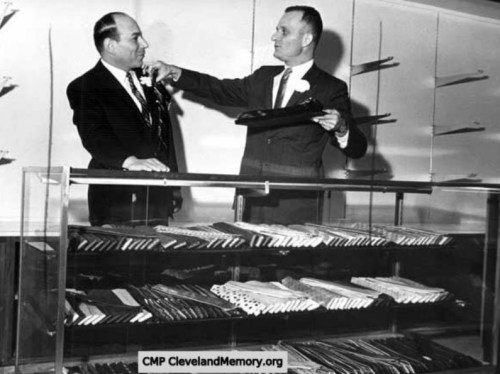 Two men in suits stand by a display case filled with various items, one man presenting something to the other.