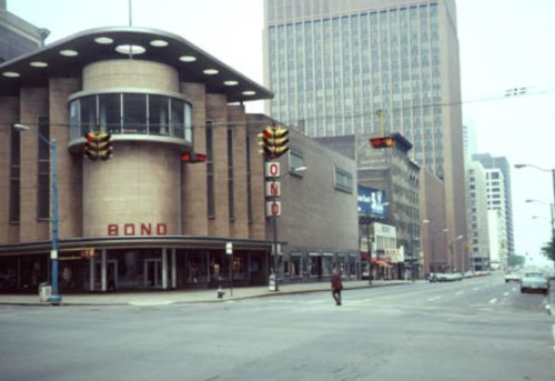A city street scene featuring a building with "BONO" signage and traffic lights, with a person walking across the road.