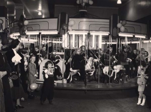 A vintage carousel with children riding, surrounded by adults and balloons in a festive indoor setting.