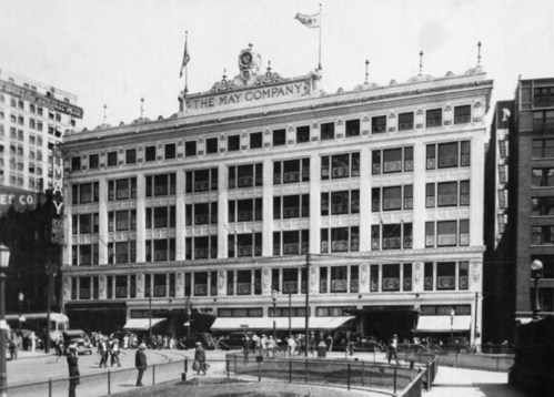 Historic black-and-white photo of The May Company building, featuring a large facade and bustling street activity.