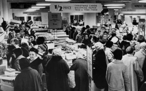 A busy store filled with shoppers browsing Christmas items, surrounded by wrapping paper and decorations.