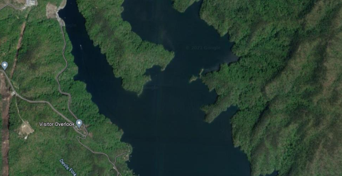 Aerial view of a lake surrounded by lush green hills and a winding road, with a visitor overlook marked nearby.