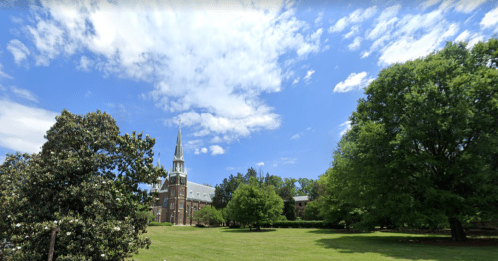 A sunny day with a blue sky over a green lawn, featuring a church with a tall steeple and surrounding trees.
