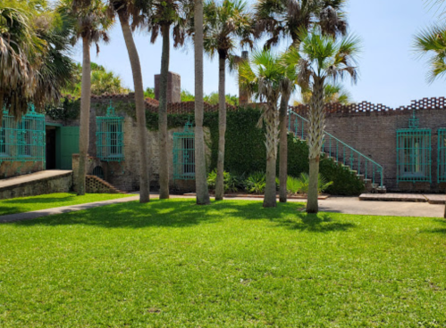 A lush green lawn surrounded by palm trees and a historic building with turquoise accents and a staircase.