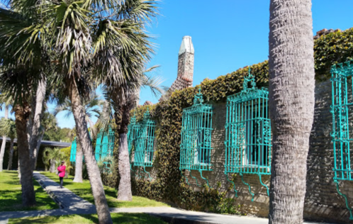 A person jogs past a stone building with turquoise window grilles, surrounded by palm trees and greenery.