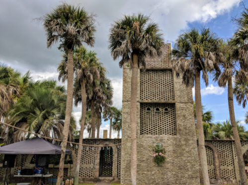 A stone building with a unique design surrounded by tall palm trees under a cloudy sky.