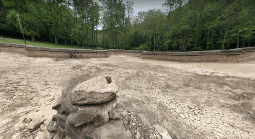 A dry, cracked landscape with a stone pile in the foreground, surrounded by lush green trees in the background.