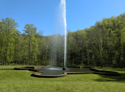 A fountain sprays water into the air, surrounded by a circular stone path and lush green trees under a clear blue sky.