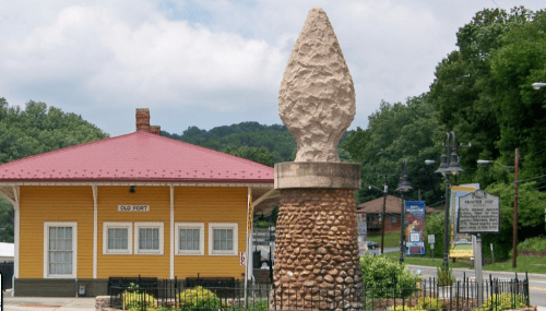 Historic yellow train station with a red roof, next to a large stone monument, surrounded by greenery and signage.