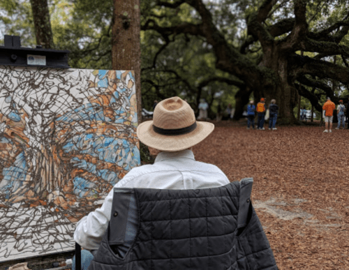 An artist in a hat sits in a chair, painting a tree scene in a park with people walking in the background.