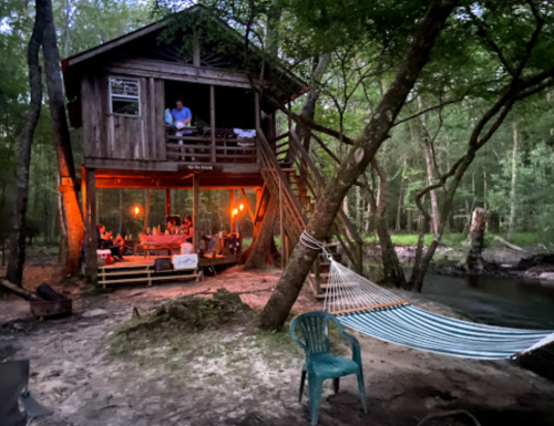 A rustic treehouse with warm lights, surrounded by trees and a hammock, near a calm stream at dusk.