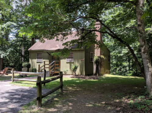 A small green cabin surrounded by trees, with a wooden porch and a gravel path leading to it.