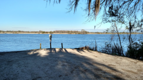 A serene river view with a sandy bank, trees, and a parking meter in the foreground under a clear blue sky.