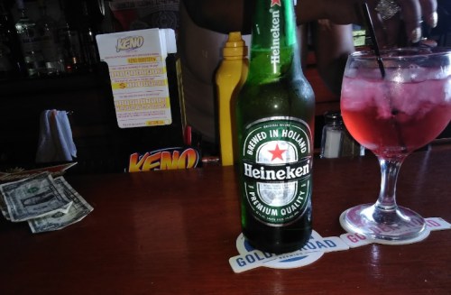A Heineken beer bottle and a cocktail in a glass sit on a bar counter with cash and a Keno game slip in the background.