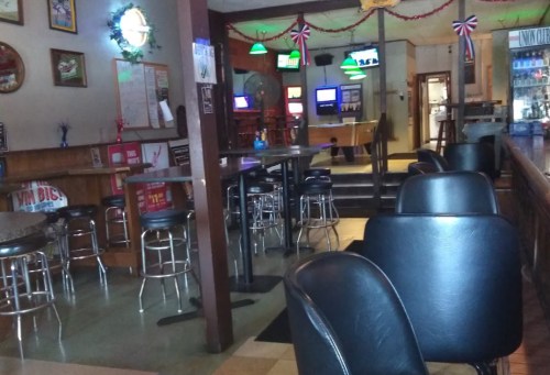 A dimly lit bar interior with empty stools, pool tables, and gaming machines in the background.